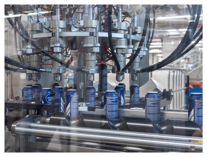 site-industries-cosmetiques Uma vista de perto de uma linha de produção industrial mostra latas de alumínio azuis a serem enchidas por uma máquina automatizada com vários bicos e tubos pretos, num ambiente industrial.