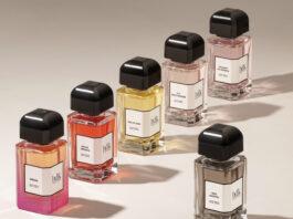 site-industries-cosmetiques Six flacons de parfum à bouchon noir sont disposés sur une surface beige. Chaque flacon contient un liquide de couleur différente et porte une étiquette blanche avec un texte noir. L'éclairage projette des ombres distinctes derrière les flacons.