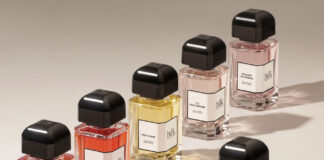 site-industries-cosmetiques 在米色表面上摆放着六个黑色瓶盖的香水瓶。每个瓶子都装有不同颜色的液体，并贴有白色标签和黑色文字。灯光在瓶子后面投射出明显的阴影。.