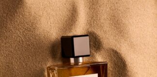 site-industries-cosmetiques 一个长方形的玻璃香水瓶，标有 Reminiscence 的 "Souffle Ambre "字样，部分被埋在浅棕色的细沙中。瓶盖为黑色，内装琥珀色液体。.