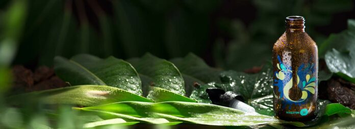 site-industries-cosmetiques A brown glass bottle with a colorful leaf pattern rests on large, wet green leaves. The bottle is covered in water droplets and surrounded by tropical foliage in natural light.