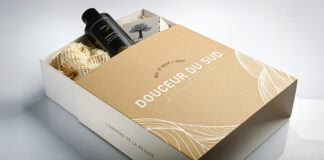 site-industries-cosmetiques Un coffret cadeau étiqueté "DOUCEUR DU SUD" contient un flacon noir, une éponge à mailles de couleur crème et une carte illustrée d'un olivier. La boîte est partiellement ouverte, révélant son contenu sur une surface blanche.