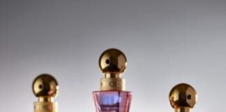 site-industries-cosmetiques Trois flacons de parfum en verre rose avec des bouchons sphériques dorés et des logos "JC" dorés sont disposés sur une surface lisse sur un fond dégradé. Les flacons sont de petite, moyenne et grande taille.