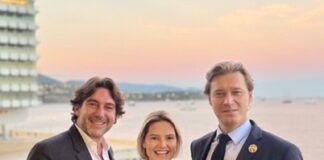 site-industries-cosmetiques Trois personnes en tenue de soirée se tiennent ensemble, souriantes, sur un balcon surplombant un plan d'eau au coucher du soleil. Un bâtiment moderne et des collines lointaines sont visibles à l'arrière-plan.