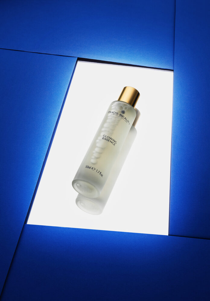site-industries-cosmetiques Un flacon givré de Biocell Smoothing Essence avec un bouchon doré est placé sur un fond blanc, entouré de panneaux géométriques bleus. Le flacon est incliné et son étiquette est visible.