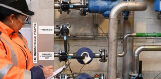 site-industries-cosmetiques Una persona con chaleco de seguridad naranja y casco escribe en un portapapeles mientras inspecciona las tuberías y válvulas industriales etiquetadas "AGUA INDUSTRIAL LIMPIA" montadas en un muro de hormigón.