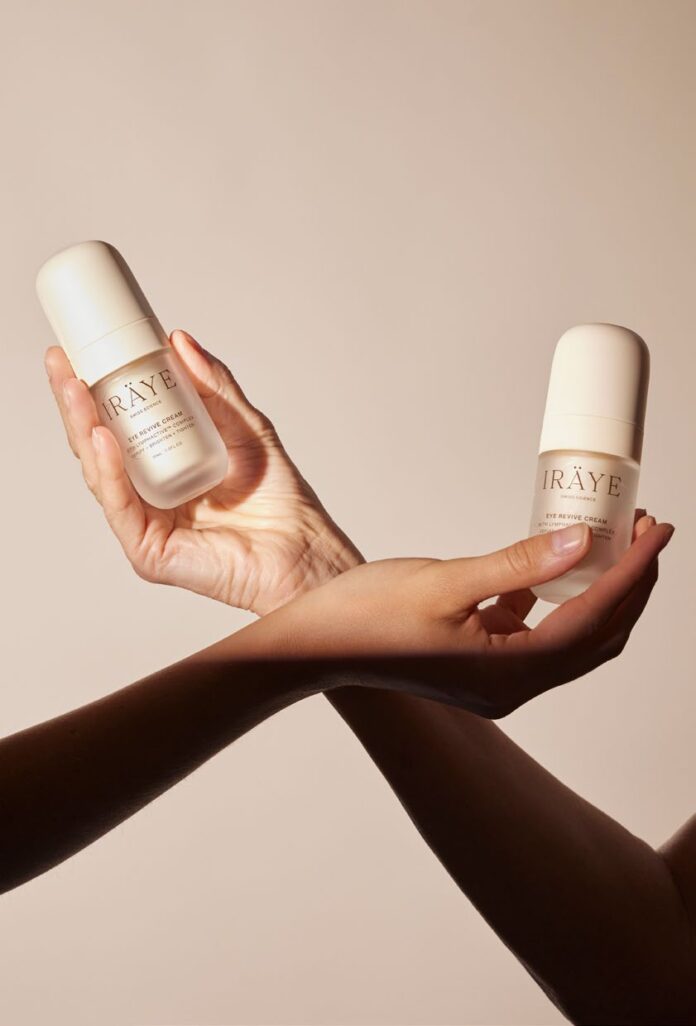 site-industries-cosmetiques 两只手拿着白色的 IRÄYE 护肤品瓶，背景是纯米色，前臂交叉在手腕处。.
