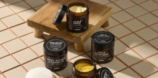 site-industries-cosmetiques Quatre pots ouverts de crèmes pour le visage Lush sont disposés sur une surface carrelée, avec deux tampons de coton et une petite plate-forme en bois à proximité. Chaque pot est étiqueté avec ses ingrédients, tels que l'avoine, le curcuma, l'huile d'argan et l'huile d'amande.