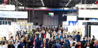 site-industries-cosmetiques Une foule de personnes se promène et se tient dans un hall d'exposition très fréquenté, avec divers stands et expositions. Beaucoup d'entre elles portent des vêtements de travail et des badges nominatifs, sous un éclairage intense.