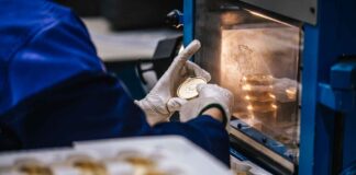 site-industries-cosmetiques Une personne portant des gants blancs et un manteau bleu place une pièce d'or ou une médaille dans une machine. D'autres pièces ou médailles sont visibles à l'intérieur de la machine et sur un plateau au premier plan.