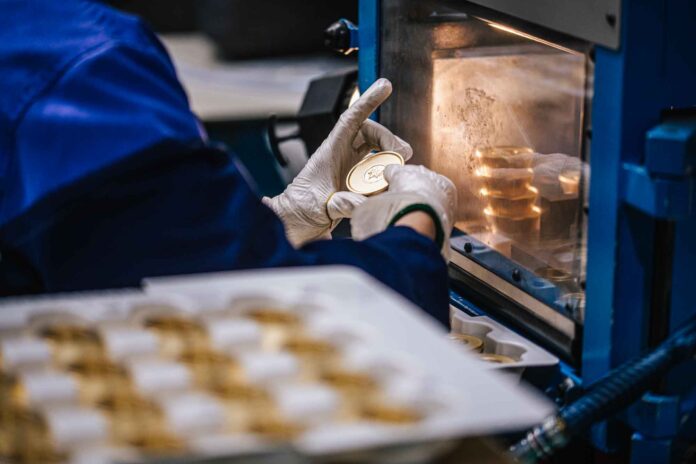 site-industries-cosmetiques 一个戴着白色手套、穿着蓝色外套的人将一枚金币或奖牌放入一台机器中。在机器内部和前景的托盘上还可以看到其他硬币或奖牌。.