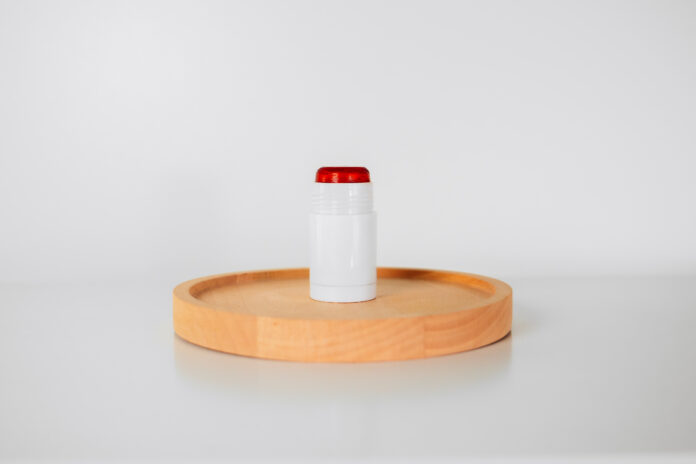 site-industries-cosmetiques Um recipiente Brouillon cilíndrico branco com uma tampa vermelha é colocado verticalmente num tabuleiro de madeira redondo sobre um fundo branco liso.