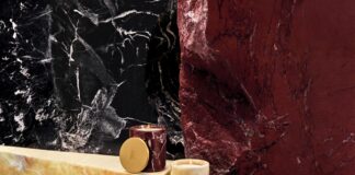 site-industries-cosmetiques Quatre bougies dans des récipients en marbre coloré sont posées sur des surfaces en pierre noire, jaune et rouge, sur un fond de marbre noir et rouge texturé. La scène met l'accent sur une esthétique luxueuse et moderne aux tons riches.