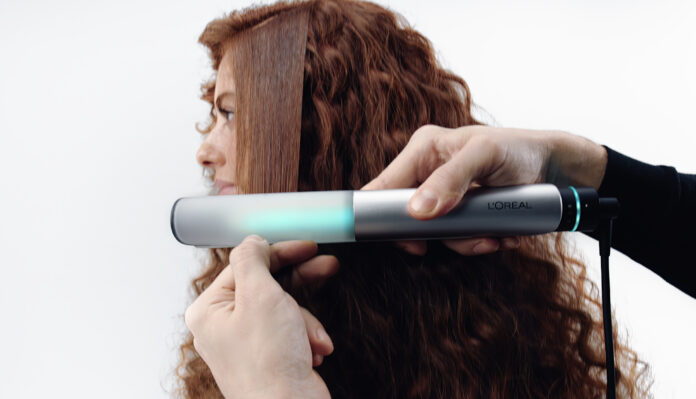 site-industries-cosmetiques Uma pessoa com cabelo ruivo comprido e encaracolado vê uma secção alisada com um aparelho de modelação da L'Oréal que emite uma luz azul-verde Auto Draft, enquanto as mãos seguram o aparelho contra o cabelo.