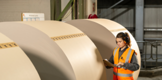 site-industries-cosmetiques Une femme portant un gilet de haute visibilité et des protections auditives examine un presse-papiers à côté de grands rouleaux de papier brun dans un entrepôt industriel.
