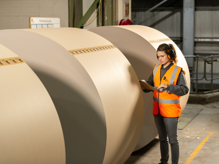 site-industries-cosmetics Uma mulher com um colete de alta visibilidade e proteção auditiva examina um pisa-papéis junto a grandes rolos de papel castanho num armazém industrial.