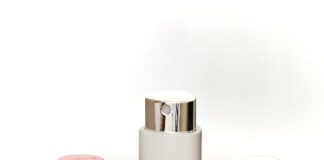 site-industries-cosmetiques Quatre flacons cylindriques de cosmétiques en spray de couleurs métalliques rose, rouge, or et blanc sont disposés verticalement et horizontalement sur une surface réfléchissante, avec un arrière-plan neutre.