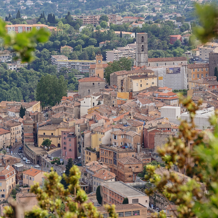 site-industries-cosmetiques Vista de uma cidade europeia com edifícios de telhado de terracota densamente povoados, uma torre de relógio e um campanário de pedra, rodeada por árvores e vegetação em segundo plano. Algumas folhas são visíveis em primeiro plano.