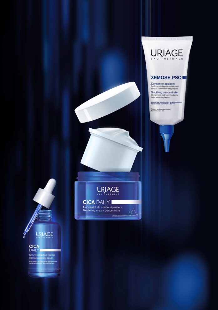 site-industries-cosmetiques Três produtos de cuidados da pele da marca Uriage estão representados num fundo abstrato azul escuro: um frasco com conta-gotas, um frasco com tampa aberta e um tubo espremido com uma ponta pontiaguda. Todos os recipientes estão rotulados com texto azul e branco.
