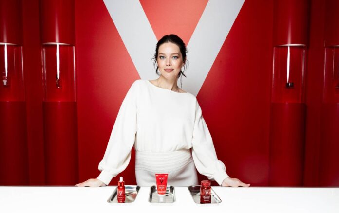 site-industries-cosmetiques Une femme vêtue d'une robe blanche se tient derrière un comptoir blanc sur lequel sont disposés trois plateaux argentés contenant des produits de soin de la peau rouges. L'arrière-plan est rouge avec un grand 