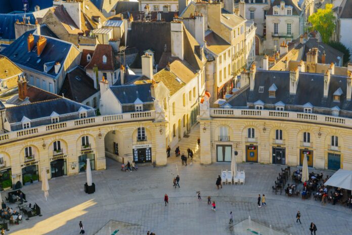 site-industries-cosmetiques Vue aérienne d'une place européenne où les gens se promènent et s'assoient à des cafés en plein air, entourés de bâtiments historiques avec des toits en pente et des cheminées, sous un soleil chaud.