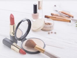site-industries-cosmetiques 白色表面上的各种化妆用品，包括两支红色口红、带刷子的粉饼、一面小巧的镜子、粉底珠、指甲油以及背景中的几支化妆刷。.