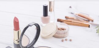 site-industries-cosmetiques Varios artículos de maquillaje sobre una superficie blanca, entre ellos dos barras de labios rojas, un polvo facial con brocha, un espejo compacto, perlas de maquillaje, laca de uñas y varios pinceles de maquillaje al fondo.