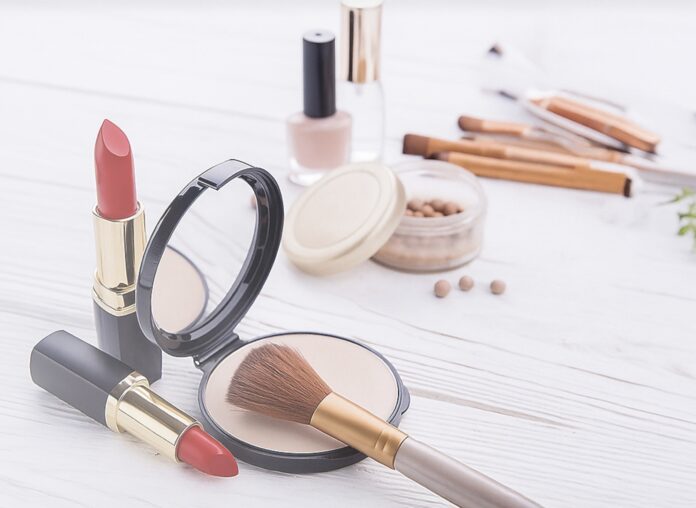 site-industries-cosmetiques Varios artículos de maquillaje sobre una superficie blanca, entre ellos dos barras de labios rojas, un polvo facial con brocha, un espejo compacto, perlas de maquillaje, laca de uñas y varios pinceles de maquillaje al fondo.