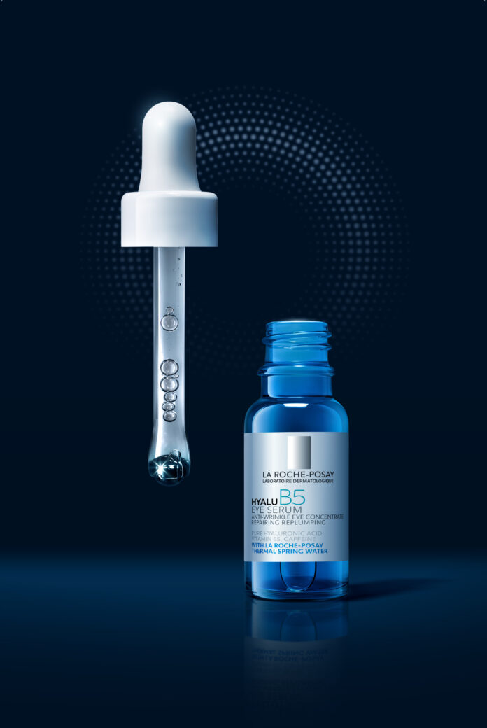 site-industries-cosmetiques Um frasco azul de La Roche-Posay Hyalu B5 Serum está ao lado de um conta-gotas com uma gota de líquido na extremidade, sobre um fundo azul escuro com um padrão circular pontilhado.