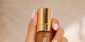site-industries-cosmetiques 一只指甲修剪成淡粉色的手，拿着一个透明瓶子，瓶子里有金色液体和金色瓶盖，瓶子上标有 "BARALAN "字样，背景是带柔和阴影的米色。.