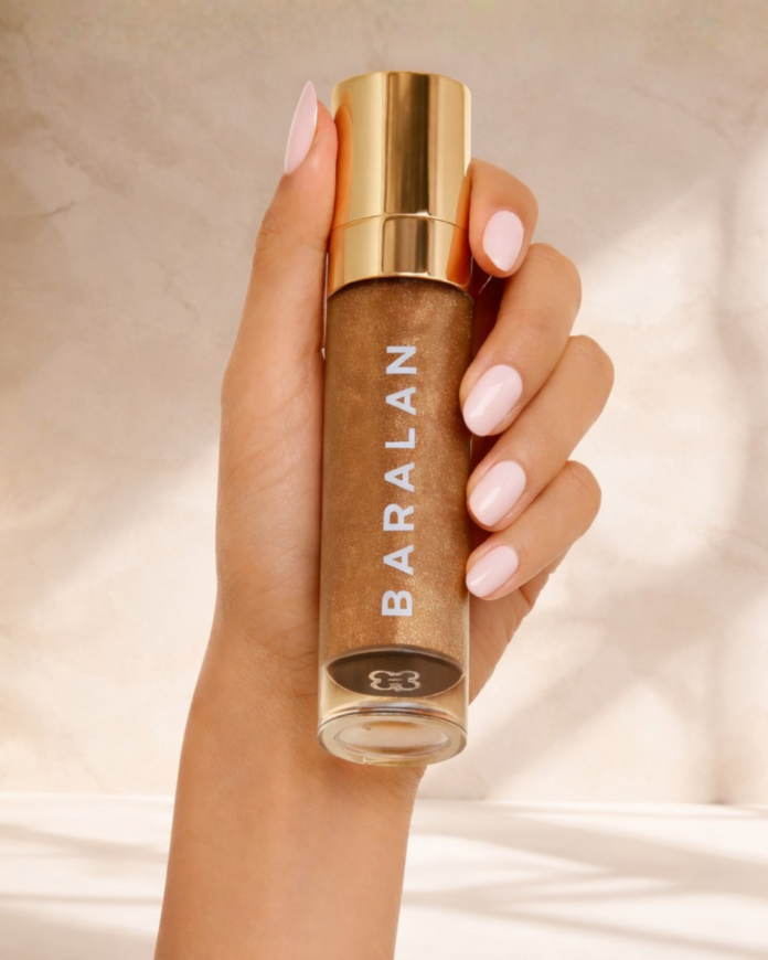 site-industries-cosmetiques Una mano con las uñas cuidadas de color rosa pálido sostiene una botella transparente con un líquido dorado y un tapón dorado, etiquetada como 