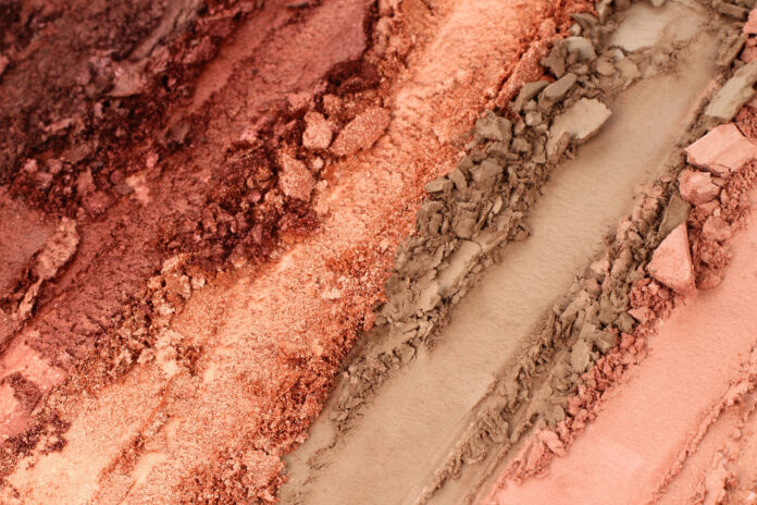 site-industries-cosmetiques Close-up of crushed make-up powders in shades of pink, peach, bronze and beige, arranged in rough, parallel strips with powdery, chunky textures.