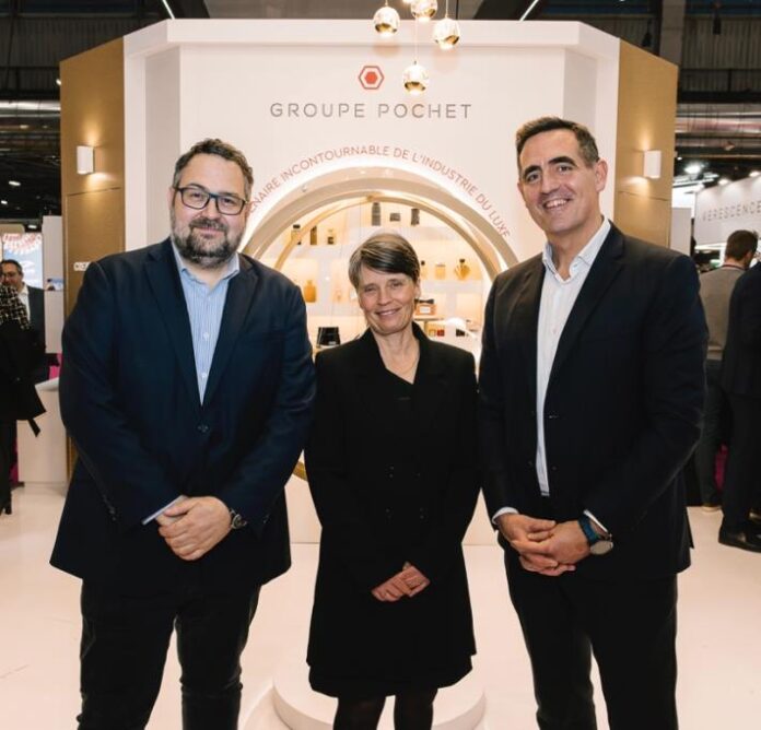 site-industries-cosmetiques Três pessoas em trajes de trabalho estão em frente a um stand do Groupe Pochet num evento em recinto fechado. O stand está a expor uma variedade de produtos e tem um motivo circular iluminado ao fundo.