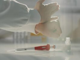 site-industries-cosmetiques 一个穿着白大褂的人在实验台上戴上乳胶手套。她面前的台子上放着一支红白相间的移液管、一个瓶子和一张纸巾。.