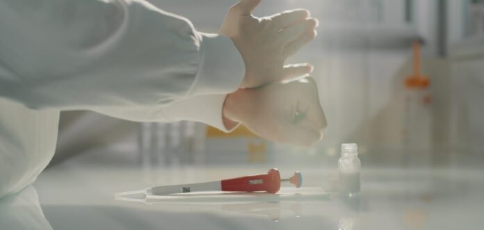 site-industries-cosmetiques Uma pessoa com uma bata de laboratório calça luvas de látex numa bancada de laboratório. Uma pipeta vermelha e branca, um frasco e uma toalha de papel estão colocados no balcão à sua frente.