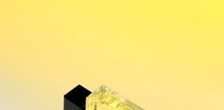 site-industries-cosmetiques 一个带黑色瓶盖的长方形玻璃香水瓶放在黄白渐变的表面上。瓶内装有黄色液体，并贴有带文字的白色标签。瓶子投下了阴影。.