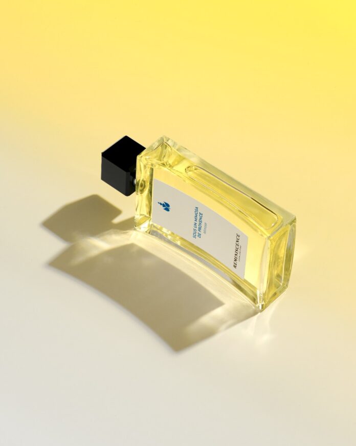 site-industries-cosmetiques Um frasco de perfume de vidro retangular com uma tampa preta assenta numa superfície com um gradiente amarelo e branco. O frasco contém um líquido amarelo e tem um rótulo branco com texto. O frasco projecta uma sombra.