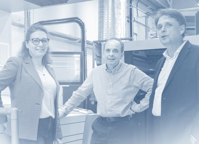 site-industries-cosmetiques Tres adultos, dos hombres y una mujer, están de pie en un entorno industrial o de oficina, con equipos y monitores visibles en el fondo. Miran hacia la cámara. La imagen está filtrada en azul.