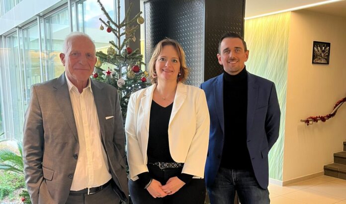 site-industries-cosmetiques Tres personas están de pie en el interior, junto a un árbol de Navidad decorado. El hombre de la izquierda lleva un traje gris, la mujer del centro lleva una americana blanca y el hombre de la derecha lleva una americana azul y cuello alto negro.