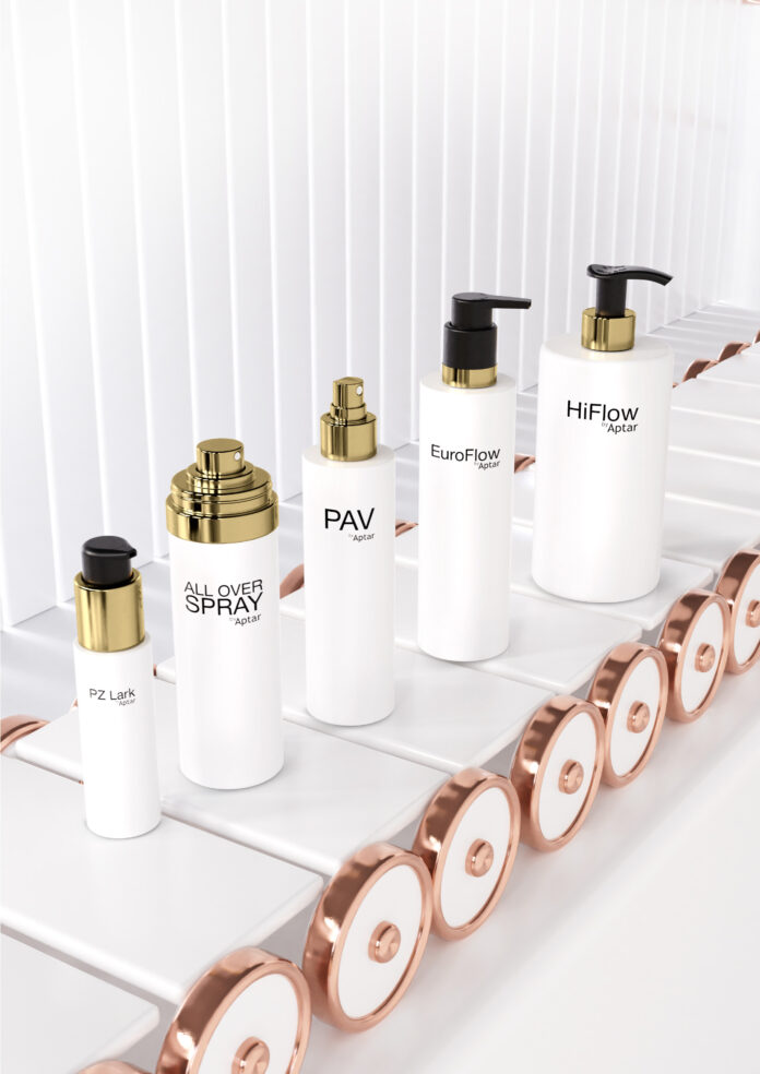 site-industries-cosmetiques Num tapete rolante branco e rosa dourado, estão expostos cinco frascos de cosméticos brancos com diferentes bombas ou sprays dourados e pretos, cada um com nomes de produtos como 