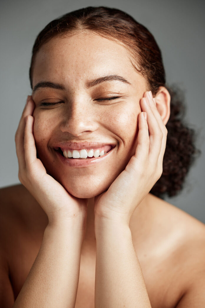 site-industries-cosmetiques A person with curly brown hair smiles, eyes closed, gently resting her face in her hands. The background is plain and gray. The person's skin appears smooth with visible freckles.