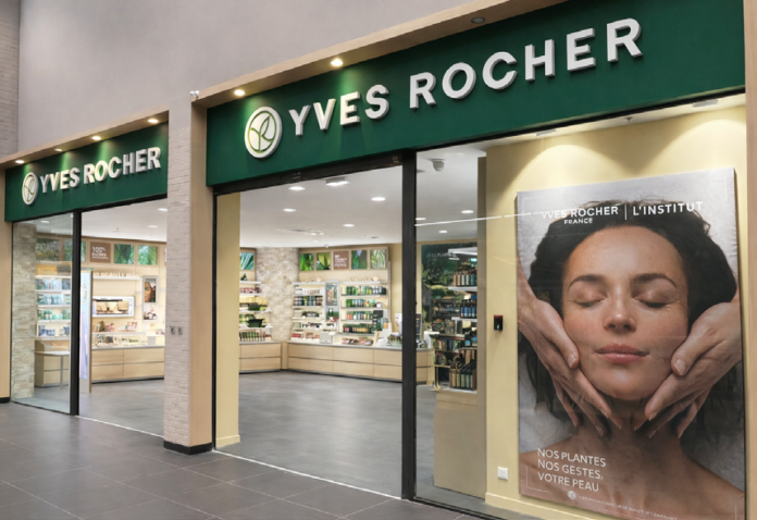 site-industries-cosmetiques Un magasin Yves Rocher avec une enseigne verte, de grandes vitrines et une affiche montrant une femme recevant un soin du visage. L'intérieur présente des étagères avec divers produits de beauté et de soins de la peau.