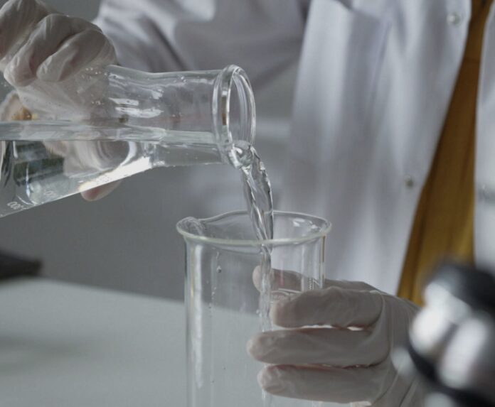 site-industries-cosmetiques Una persona con bata blanca y guantes vierte un líquido transparente de un matraz a un vaso de precipitados sobre una mesa de laboratorio.