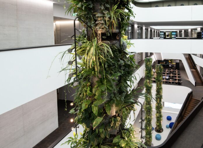 site-industries-cosmetiques L'intérieur moderne à plusieurs niveaux se caractérise par de hautes colonnes couvertes de plantes vertes, des murs blancs, un sol sombre, des allées ouvertes et des zones de sièges avec des chaises bleues.