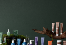 site-industries-cosmetiques Une variété de produits de soins de la peau dans des emballages colorés sont disposés sur des surfaces couvertes de mousse avec de petites fleurs et un morceau de bois flotté sur un fond vert foncé.