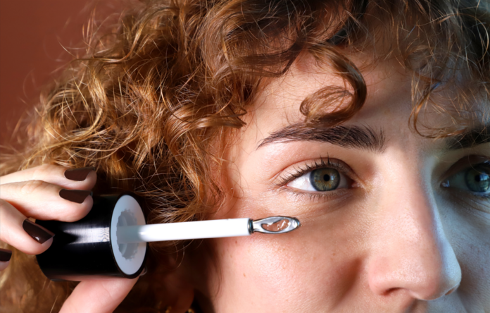 site-industries-cosmetiques Una persona con el pelo rizado sostiene una varita de rímel cerca de su ojo derecho, preparándose para aplicar el producto. Su rostro se muestra en primer plano, centrándose en el ojo y la ceja.
