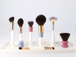 site-industries-cosmetiques 一套六支形状和大小各异的化妆刷，带白色手柄和金色装饰，摆放在一个白色托盘上。托盘前摆放着两把双头化妆刷。.