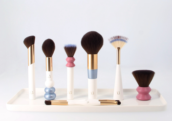 site-industries-cosmetiques Conjunto de seis pincéis de maquilhagem de várias formas e tamanhos, com cabos brancos e detalhes dourados, dispostos num tabuleiro branco. Dois pincéis de ponta dupla estão colocados em frente ao tabuleiro.