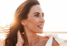site-industries-cosmetiques Une femme aux longs cheveux bruns regarde sur le côté et sourit, debout à l'extérieur, sous la lumière du soleil. Elle porte un haut blanc avec un nœud sur l'épaule. L'arrière-plan est lumineux et semble être une plage ou un paysage ouvert.