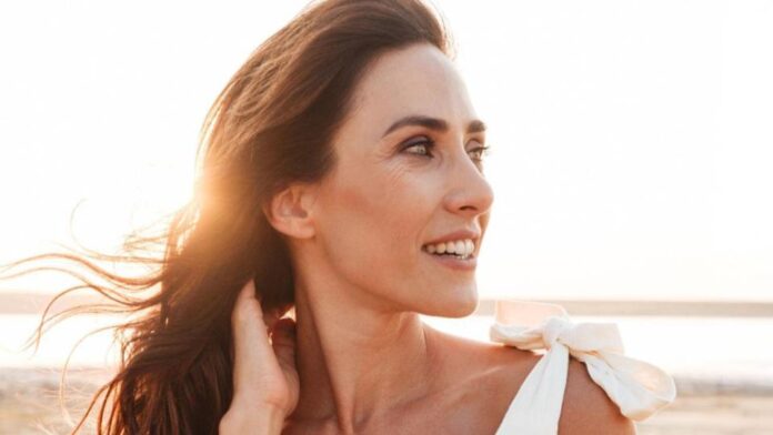 site-industries-cosmetiques Una mujer con el pelo largo y castaño mira a un lado y sonríe, de pie al aire libre, bajo la luz del sol. Lleva un top blanco con un lazo en el hombro. El fondo es luminoso y parece una playa o un paisaje abierto.